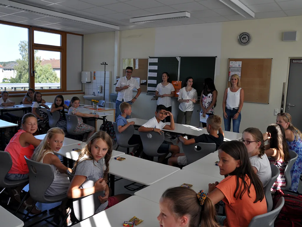 Kinder sitzen im Klassenraum, eine Lehrkraft steht bei ihnen.